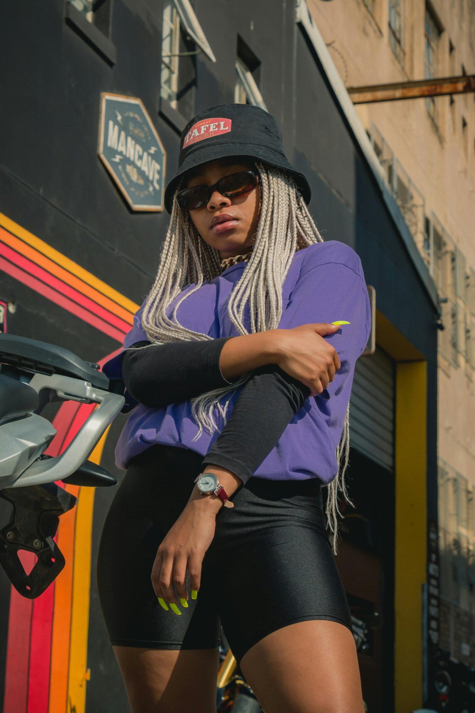 Fashionable woman with braids, sunglasses, and bucket hat posing in Cape Town urban setting.